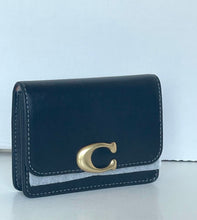 Load image into Gallery viewer, A black leather wallet with a prominent gold-tone 'C' logo clasp on the front, resting on a light gray surface against a white background.