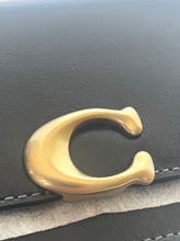 Load image into Gallery viewer, Close-up of a gold-tone metal Coach 'C' logo clasp attached to a black leather strap, with a textured light gray fabric visible underneath.
