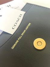 Load image into Gallery viewer, Close-up of a Coach wallet or card holder in black full-grain leather with gold text reading 'GENUINE FULL GRAIN LEATHER' and a gold snap closure, with a white Coach tag partially visible.
