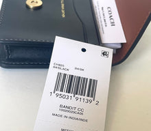 Load image into Gallery viewer, A white COACH product hangtag with barcode and pricing details placed over a dark brown leather wallet on a light beige surface.