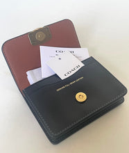 Load image into Gallery viewer, An open Coach full-grain leather wallet in black and brown, showing card slots, a gold snap button, and branded paper tags on a light neutral background.