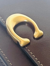 Load image into Gallery viewer, Close-up of a polished gold-tone metal C-shaped hardware piece attached to dark brown leather trim with white stitching.
