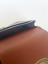 Load image into Gallery viewer, Close-up detail of a brown leather handbag showing the edge of a black strap and a gold-tone metal clasp against a plain light background.