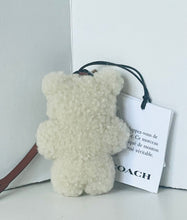 Load image into Gallery viewer, A cream-colored teddy bear-shaped fluffy accessory with a black strap and a white COACH brand tag, resting on a white surface against a white background.