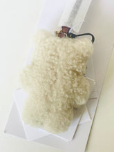 Load image into Gallery viewer, A cream-colored, fluffy sheepskin or faux-shearling pouch with a small brown leather strap and black zipper pull, resting on white geometric paper packaging.