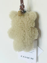 Load image into Gallery viewer, A Coach teddy bear-shaped fluffy cream bag charm with a brown leather strap and a gold-tone metal logo tag, shown with its branded hangtag against a plain white background.
