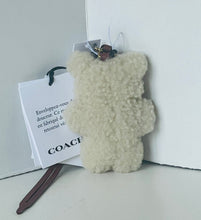 Load image into Gallery viewer, A cream-colored, textured teddy bear-shaped keychain or bag charm with a dark brown leather strap and a white paper tag printed with Coach branding, set against a minimalist white background.