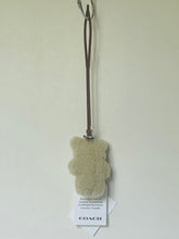 Load image into Gallery viewer, A Coach teddy bear-shaped bath bomb in a light beige textured material, hanging from a brown strap with its white retail tag below, against a plain off-white wall.