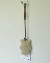 Load image into Gallery viewer, A Coach teddy bear-shaped bath sponge in a light beige bouclé texture, hanging from a brown leather strap on a metal wall hook. A white paper tag with the Coach logo is attached at the bottom.