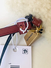 Load image into Gallery viewer, A Coach leather accessory tag with a gold-tone metal tag and a brown leather strap, attached to a white retail hangtag with the Coach logo, set against a cream-colored textured background.