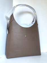 Load image into Gallery viewer, A taupe brown leather shoulder bag with a curved top handle, partially covered with protective plastic wrapping. The bag features a front pocket with a small gold button detail and a hangtag on the side, set against a plain white background.