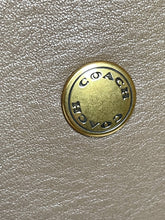 Load image into Gallery viewer, A close-up of a round gold-tone metal button engraved with the brand name 'COACH' in reverse, set against a textured beige leather background.