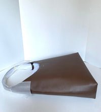 Load image into Gallery viewer, A brown leather shoulder bag with a long strap, still partially wrapped in protective plastic, resting on a white surface against a white background.