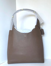 Load image into Gallery viewer, A Coach hobo shoulder bag in taupe brown leather, still wrapped in protective plastic on the handle, with a small brand tag hanging from the side.