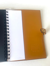 Load image into Gallery viewer, A tan leather spiral notebook cover holding an open black spiral notebook with blank white pages, photographed on a light neutral background.