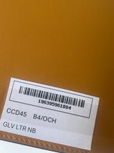 Load image into Gallery viewer, A close-up of a tan textured leather surface with a white product tag attached, showing a barcode and printed text including 'GLV LTR NB'.