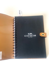 Load image into Gallery viewer, A Coach spiral-bound notebook or planner shown open, with a black front cover featuring the white COACH logo and a tan interior cover, photographed on a plain light background.