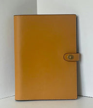 Load image into Gallery viewer, A tan leather planner cover with a snap-button strap closure, standing upright against a white corner backdrop.
