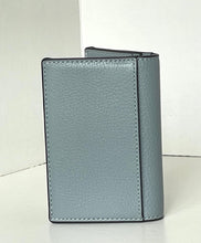 Load image into Gallery viewer, A light blue-gray pebbled leather bifold wallet standing upright, showing a black zipper along the top edge, set against a plain off-white background.