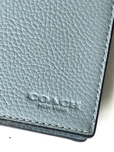 Load image into Gallery viewer, Close-up of a Coach New York pebbled leather card holder or wallet in a muted blue-gray color, showing the debossed brand name and white contrast stitching.