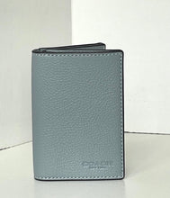 Load image into Gallery viewer, A Coach slim bifold wallet in textured light blue leather with black trim and white stitching, standing upright on a white surface against a white background.