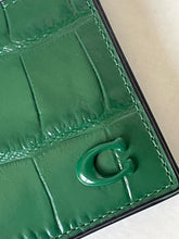 Load image into Gallery viewer, Close-up of a green crocodile-embossed leather item with a glossy green monogram letter 'C' clasp and black border on a light neutral background.