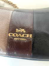 Load image into Gallery viewer, Close-up of a Coach New York handbag showing a dark brown and black leather body with a gold-tone metal logo plate featuring the signature horse and carriage emblem.