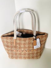 Load image into Gallery viewer, A Coach woven straw tote bag with light beige fabric handles, featuring the Coach logo embroidered in tan on the front and attached product tags.