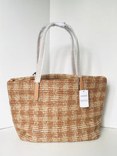 Load image into Gallery viewer, A woven straw tote bag with a natural tan and beige pattern, featuring light gray fabric handles with tan leather accents and a white hangtag.