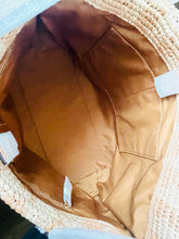 Load image into Gallery viewer, Close-up of the interior of a woven straw or raffia handbag, showing a tan fabric lining with a front slip pocket and small white tags attached.