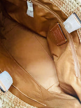 Load image into Gallery viewer, Close-up of the interior of a woven straw tote bag, showing a tan satin lining, zippered compartments, and a small brown leather brand patch.
