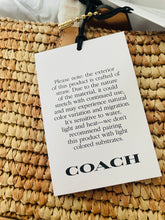 Load image into Gallery viewer, A close-up of a woven straw bag with a white COACH hangtag attached, showing the brand name and care instructions printed on the tag.