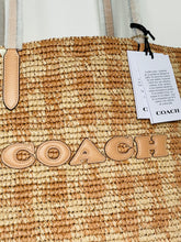 Load image into Gallery viewer, A close-up of a Coach woven straw tote bag with tan leather lettering spelling COACH across the front and a white product tag hanging from the handle.