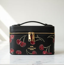 Load image into Gallery viewer, A Coach top-handle cosmetic case in black faux leather with a red cherry sequin floral pattern and gold-tone hardware, shown on a neutral beige background.