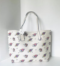 Load image into Gallery viewer, An off-white tote bag with a repeating pattern of colorful cartoon dogs, featuring light gray handles and a hanging white brand tag.