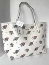 Load image into Gallery viewer, A Coach tote bag in off-white coated canvas with an allover print of colorful cartoon cats and a silver-tone metal logo plate, shown with its retail hangtag against a neutral gray background.