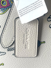 Load image into Gallery viewer, A close-up of a Coach New York leather keychain tag in white textured leather with a silver-tone metal border, attached to a white branded hangtag. The Coach logo is embossed on the tag, and part of a patterned strap is visible in the background.