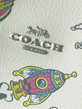 Load image into Gallery viewer, Close-up of a Coach New York logo in silver metal on a textured off-white leather surface with colorful cartoon rocket graphics.