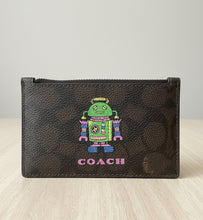 Load image into Gallery viewer, A Coach zip-around pouch in dark brown textured leather with a colorful embroidered robot design and the word 'COACH' in pink below it, shown against a white background.