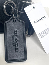 Load image into Gallery viewer, A black textured leather Coach keychain tag with an embossed vertical COACH logo, attached to a metal key ring and shown with a white COACH hangtag on a light surface.