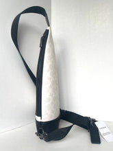 Load image into Gallery viewer, A Coach crossbody bag in white and black monogram canvas, shown standing upright against a white wall with its black strap draped to the side and a hangtag visible.