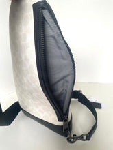 Load image into Gallery viewer, A close-up of a shoulder bag showing the side profile with the main zip open, revealing a dark gray interior lining. The exterior has a light beige and white monogram pattern with black trim and a black adjustable strap attached.