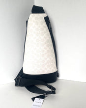Load image into Gallery viewer, A Coach sling bag in their signature monogram canvas, featuring a cream and beige pattern with black trim and a black adjustable shoulder strap. A white brand tag is attached to the strap, and the bag is photographed standing on a white surface against a white background.