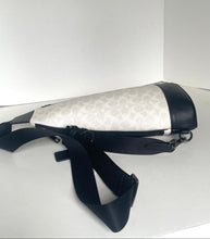 Load image into Gallery viewer, A Coach crossbody bag in white and black monogram canvas with black leather trim and a wide black strap, displayed on a white surface against a white background.