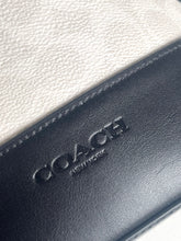 Load image into Gallery viewer, Close-up of a Coach New York leather accessory showing a black embossed logo on a dark leather section, with a light cream textured leather surface in the background.