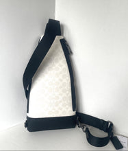 Load image into Gallery viewer, A Coach sling bag in black and off-white monogram canvas, shown standing in a white corner with its detachable strap laid out.
