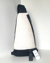 Load image into Gallery viewer, A Coach sling bag in cream and black monogram canvas, shown standing upright in a white corner. The bag features a black shoulder strap, black base, and black zipper, with a black keychain and white product tags hanging from the zipper.