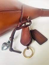 Load image into Gallery viewer, Brown leather keychain with heart charm, gold ring, and Coach logo tag attached to a brown leather bag.