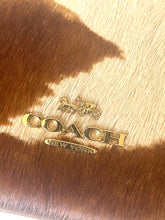Load image into Gallery viewer, Close-up of a Coach New York logo in gold-tone metal on a brown and beige cowhide-textured surface.