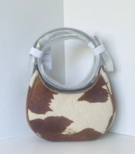 Load image into Gallery viewer, A Coach houndstooth shoulder bag in brown and cream cowhide, featuring a clear plastic-wrapped circular handle and a silver zipper closure, set against a white corner background.